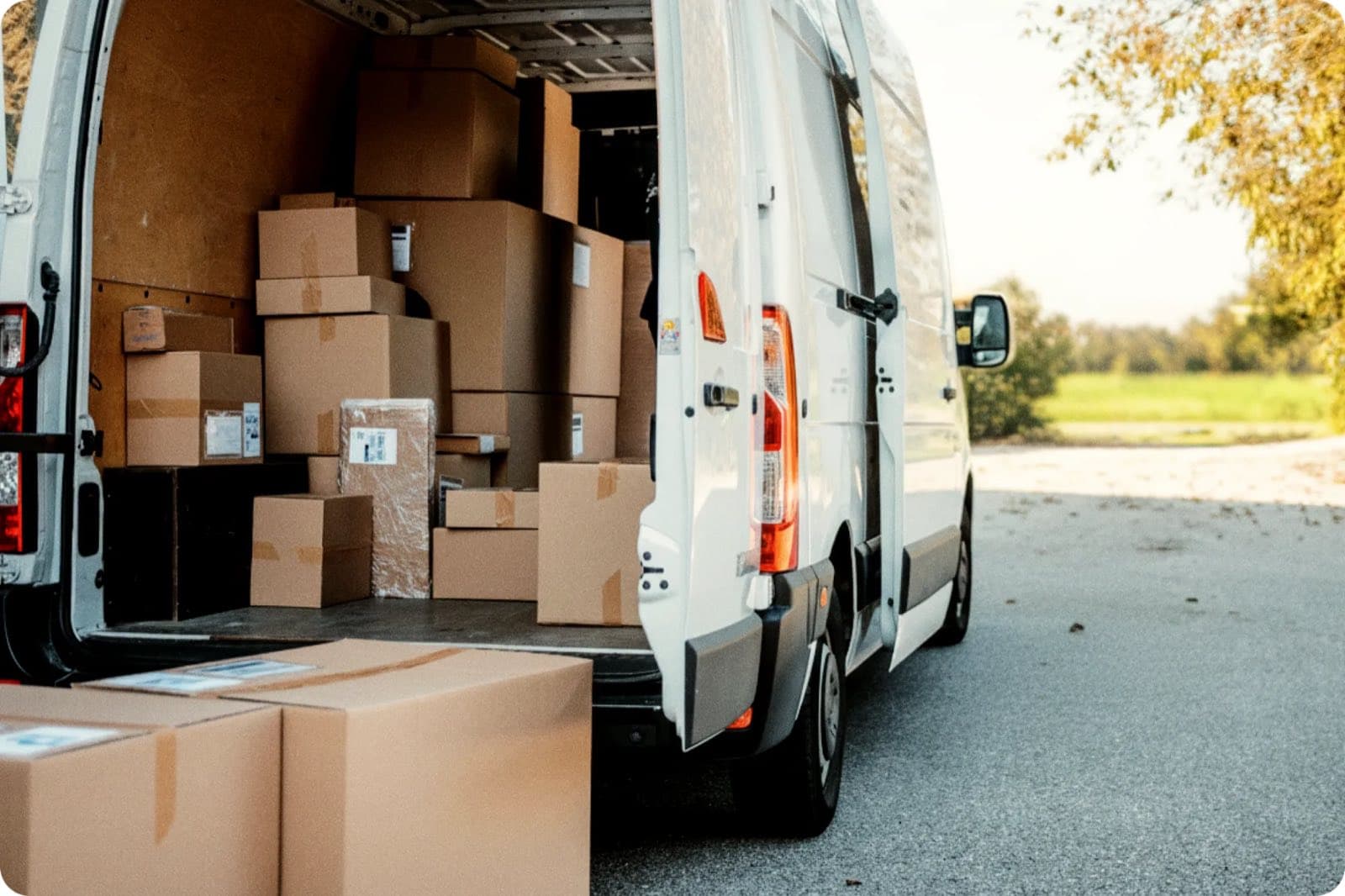The open rear doors of a white cargo van packed to the ceiling with stacked cardboard boxes and packages, parked outside on a paved driveway