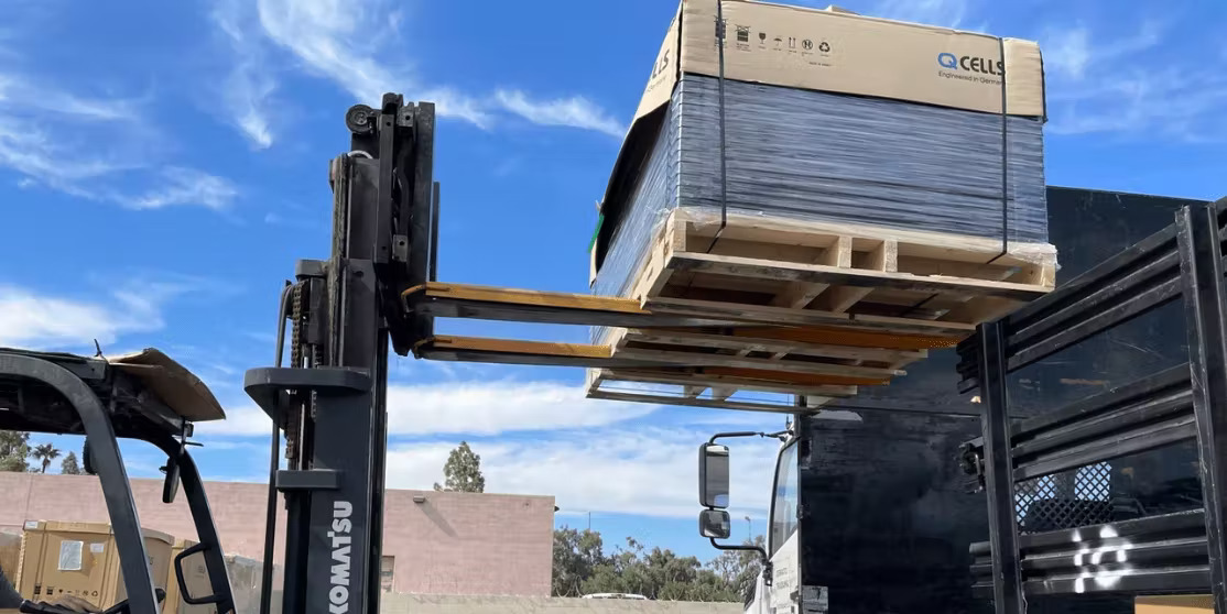 A forklift is lifting a large pallet of boxed solar panels onto a flatbed truck.