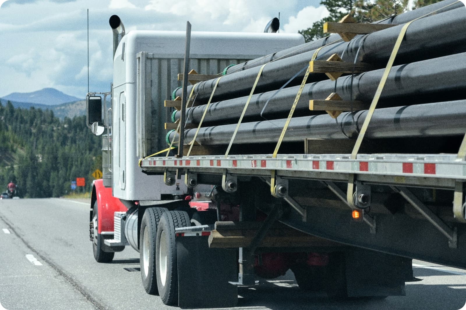 A flatbed truck hauling large industrial pipe secured with straps and wood blocking, driving along a mountain highway
