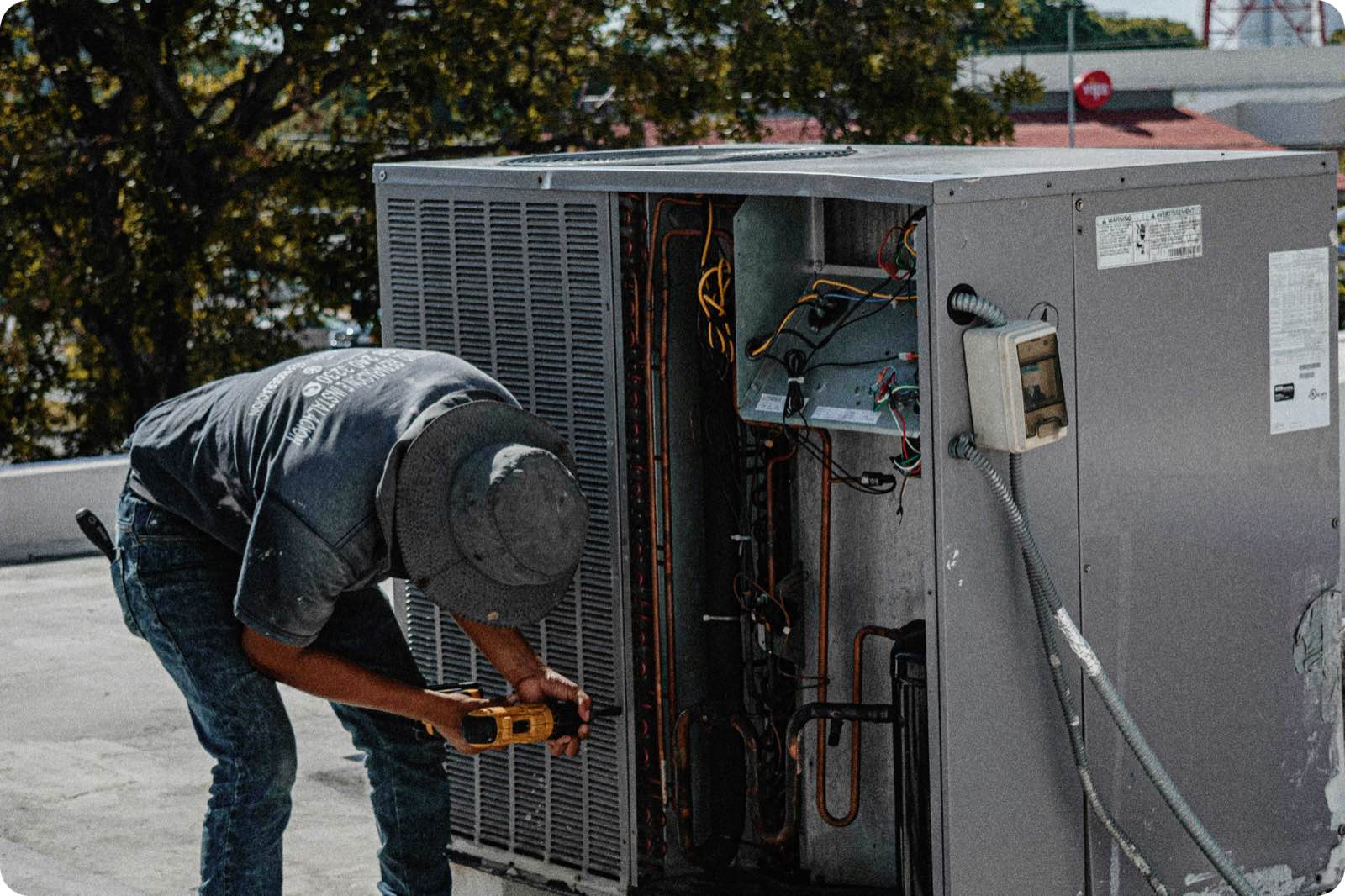 An HVAC technician in a wide-brimmed hat kneeling to service an outdoor rooftop condensing unit with a power drill, wiring and copper refrigerant lines exposed inside the open panel