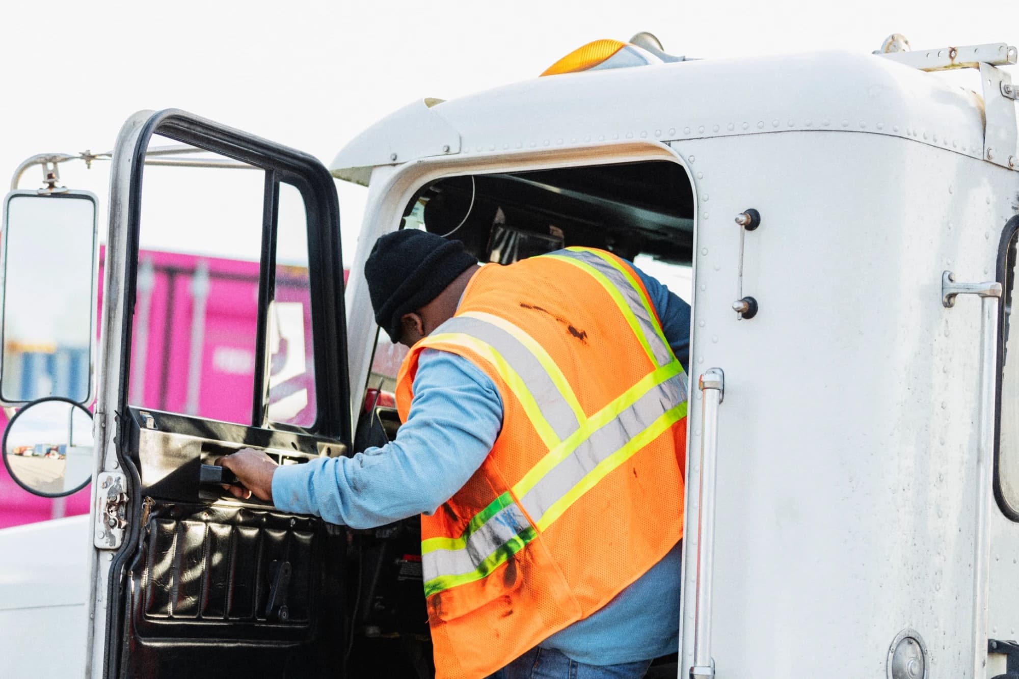 A truck driver in an orange high-visibility safety vest and knit cap climbs into the cab of a white semi-truck, gripping the open door